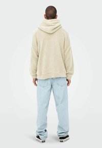 Cremfarbener Fleece-Hoodie mit strukturierter Außenseite, oversize Schnitt und Kordelzugkapuze. Kombiniert mit hellblauen Jeans und schwarzen Sneakers.