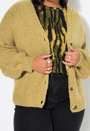 Personne en train d'attacher les boutons d'un cardigan en grosse maille couleur moutarde porté sur un haut imprimé animal noir et jaune avec un pantalon noir.
