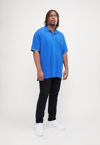Modrá polo tričko s krátkými rukávy a špičatým límcem, spárované s černými slim-fit kalhotami a bílými teniskami.