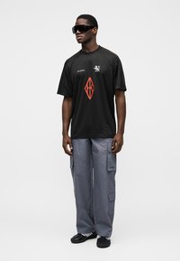 Μαύρο κοντομάνικο t-shirt με κόκκινο γεωμετρικό σχέδιο και λευκό λογότυπο. Ανοιχτό γκρι παντελόνι cargo με τσέπες. Μαύρα αθλητικά παπούτσια και γυαλιά ηλίου.