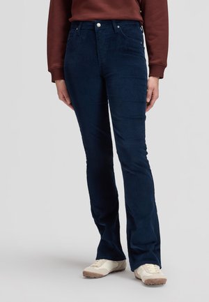 Personne portant un pantalon en velours côtelé bleu foncé, un haut à manches longues marron et des baskets blanches, debout sur un sol gris clair uni.