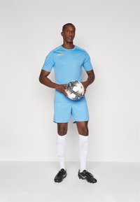 Lichtblauw voetbaluniform met korte mouwen, bijpassende shorts, witte sokken met kniebeschermers en zwarte voetbalschoenen. De speler houdt een zwart-witte voetbal vast.