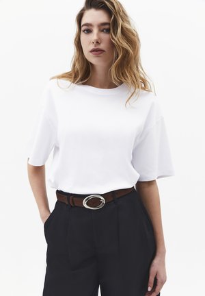 Kobieta o falowanych blond włosach nosi prostą białą koszulkę z krótkim rękawem włożoną do czarnych spodni z brązowym paskiem i srebrną klamrą.