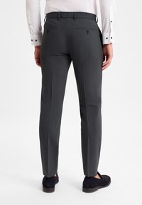 Graue maßgeschneiderte Hosen mit einem strukturierten Muster, ausgestattet mit zwei Gesäßtaschen mit Knopfverschluss, kombiniert mit dunklen Slippers.