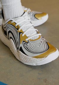 Witte sportschoen met zwarte en gouden accenten. Heeft een golvend patroon en een textuur op de witte zool. Veters zijn stevig gestrikt.