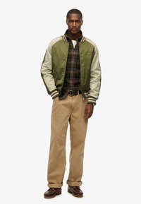 Grøn og beige bomberjakke med stribede manchetter, lagdelt over en ternet skjorte. Beige bukser og brune sko fuldender outfittet.