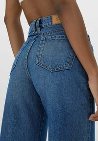 Korkeavyötäröiset siniset farkut rentoa istuvuutta, joissa on takataskut ja nahkaleima vyötäröllä. Pehmeä tekstuuri ja klassinen muotoilu.