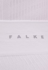 Białe skarpetki dziane z ribbingu, z szarym logo "FALKE" blisko góry, gładkim paskiem i trójkątnymi wzorami na cholewce.