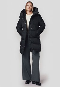 Czarna pikowana kurtka z kapturem; posiada napy i kieszenie. Noszona na lekkim swetrze i szarych szerokich spodniach. Gładka struktura.