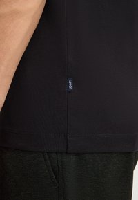 Zwarte kortekorte t-shirt van gladde stof, voorzien van een klein marineblauw label met de merknaam aan de zijkant; gecombineerd met donkergroene gestructureerde broek.