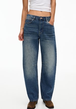 Femme portant un jean bleu délavé ample avec des poches avant, associé à des bottines marron et un top court blanc, se tenant avec une main dans la poche.