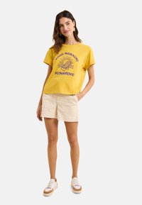 T-shirt en coton jaune avec un graphique "BONJOUR SOLEIL" en violet, associée à un short beige. Modèle portant des baskets blanches.