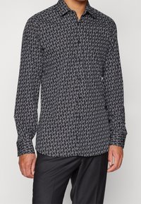 Camisa de manga larga en patrón blanco y negro con cuello, que presenta un diseño abstracto repetitivo sobre una tela ligera. Puños con botones.