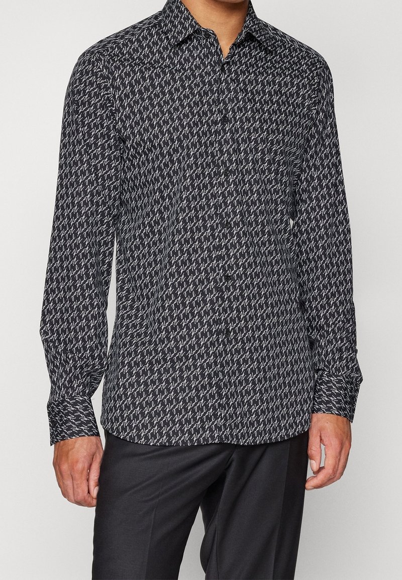 Camisa de manga larga en patrón blanco y negro con cuello, que presenta un diseño abstracto repetitivo sobre una tela ligera. Puños con botones.