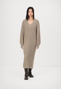 Beige stickad klänning med V-ringning och långa, puffärmar. Den har en ribbad textur, når till mitt på vaden och är ihop med mörka ankelboots.