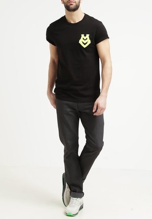 Mand iført sort t-shirt med gul logo på brystet, mørke bukser og hvide sneakers med grønne detaljer, stående mod en hvid baggrund.