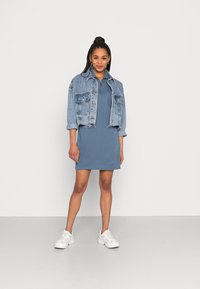 Casaco de ganga com dois bolsos frontais, comprimento curto, sobre um vestido azul sólido e curto. Modelo veste ténis brancos, luz natural, fundo simples.