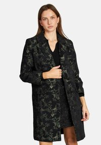 Mujer con cabello castaño lacio que lleva un abrigo largo negro con un patrón floral sobre una blusa negra de escote en V y una falda negra texturizada.