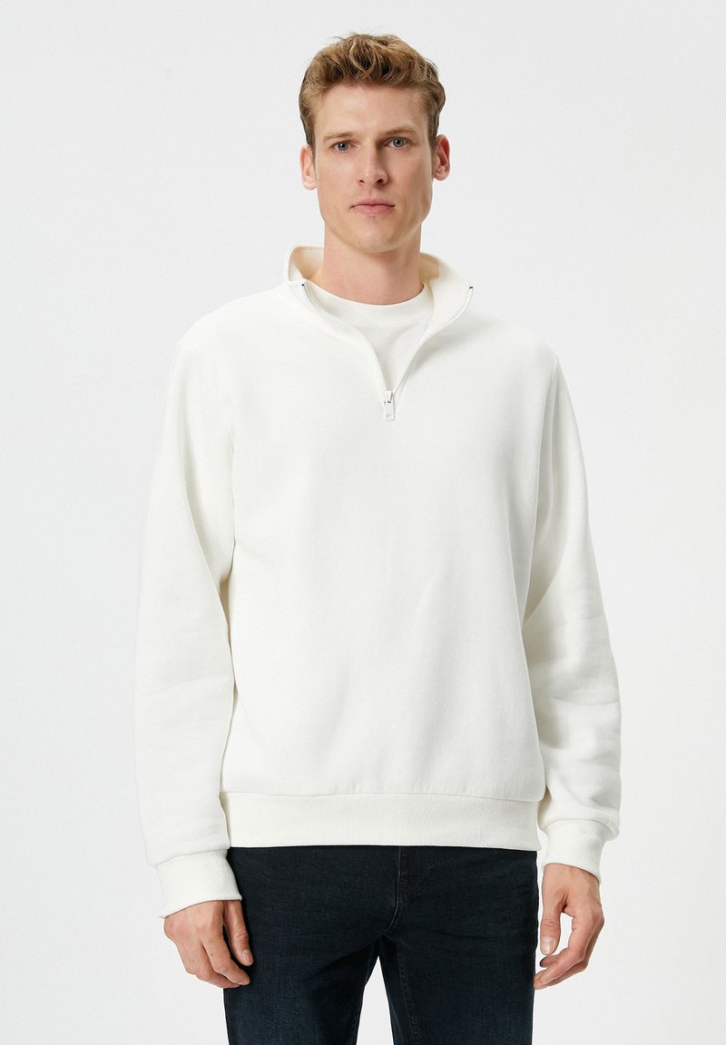 Man in een witte kwart-rits pullover met geribbelde mouwen en zoom, gecombineerd met donkere jeans, staande tegen een plain witte achtergrond.