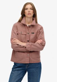 Różowa bawełniana kurtka z guzikami z przodu, dwoma kieszeniami na piersi i z kołnierzem opadającym, stylizowana z niebieskimi dżinsami na casualowy wygląd.