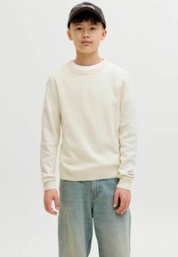 Jeune personne portant un pull crème, un jean bleu clair et une casquette noire, debout devant un fond clair uni.