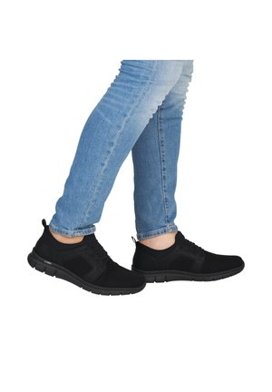 Füße in schwarzen Netz-Sneakern und blauen Jeans, mit einem leicht angehobenen Bein, vor weißem Hintergrund.