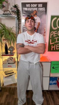 Camiseta blanca con gráfico rojo, pantalones de chándal grises y gafas de sol sobre la cabeza. Las características visuales clave incluyen un estilo de ropa deportiva casual y un ajuste relajado.
