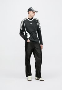 Fekete sportos póló fehér részletekkel és három csíkkal az ujjakon, sötét farmernadrággal és fehér tornacipővel párosítva. A kinézetet sapka egészíti ki.