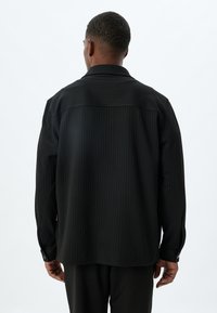 Camisa negra de manga larga con rayas verticales, puños abotonados y un cuello clásico. El material parece suave y ligeramente texturizado.