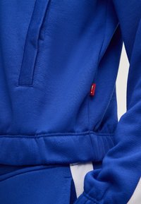 Sweatshirt blu in tessuto strutturato, con apertura per una tasca laterale e un piccolo etichetta rossa sulla cucitura. Polsi elasticizzati e vita elastica.