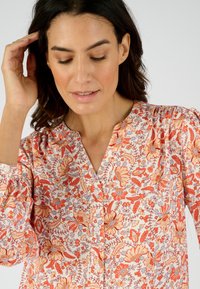 Bloes met bloemenpatroon in het wit met oranje en blauwe patronen, voorzien van een V-hals, geplooide details en lange mouwen. Gestructureerde stof.