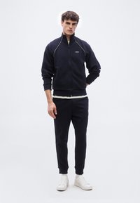 Νέος άνδρας που φοράει σκούρο μπλε φόρμα με ζακέτα με φερμουάρ και λευκά αθλητικά παπούτσια, στέκεται μπροστά σε απλό φωτεινό φόντο.