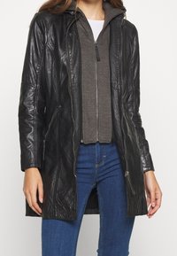Schwarze Lederjacke mit Kapuze, ausgestattet mit Reißverschlusstaschen und einer glatten Textur, über einem grauen Reißverschluss-Hoodie und blauen Jeans getragen.