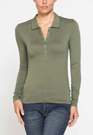 Donna che indossa una polo verde oliva a maniche lunghe con bottoni e jeans blu, mostrata dalle spalle ai fianchi.