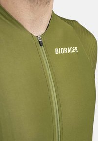 Maillot de cyclisme vert olive avec une fermeture éclair à l'avant, présentant une texture côtelée et le logo "BIORACER" en blanc sur le côté gauche.