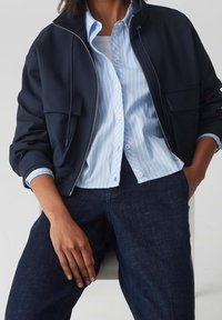 Marinblå bomberjacka med dragkedja och två framfickor, över en ljusblå randig skjorta med knäppning och mörk denimjeans.