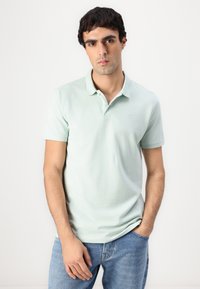 Lys grønn polo med klassisk krage, korte ermer, og subtilt brodert logo på brystet. Brukt med blå jeans.