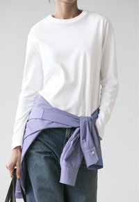 Person wearing a white long-sleeve shirt with a blue striped shirt tied around waist and dark denim jeans, holding a brown bag.