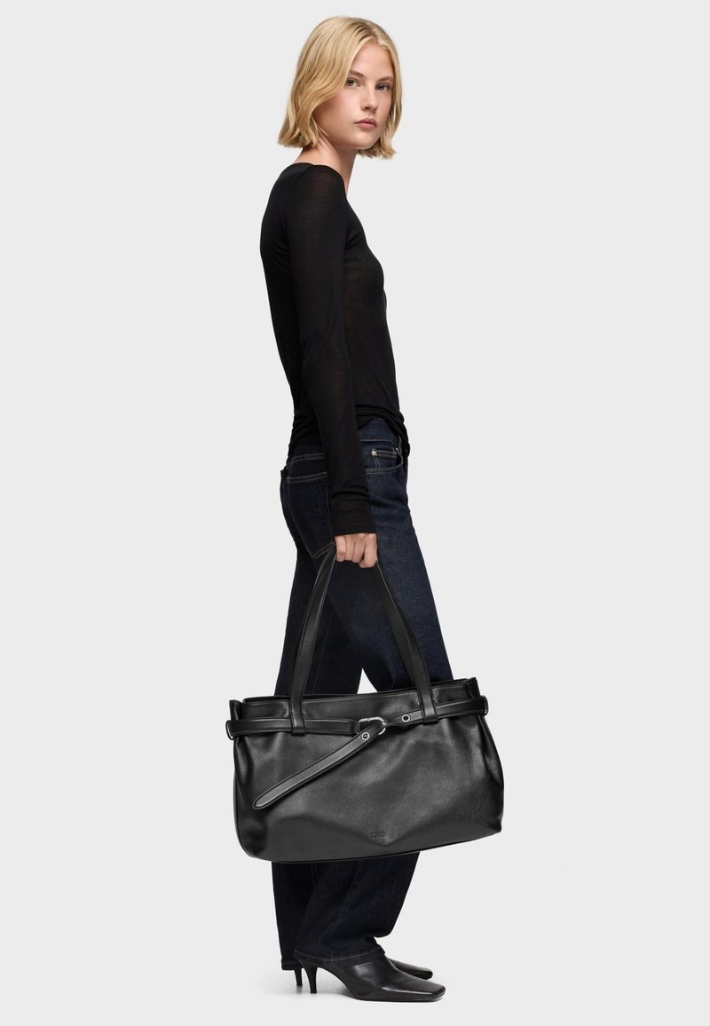 Borsa tote in pelle nera con doppi manici, texture liscia, tracolla regolabile e hardware argentato. Modella con top nero e jeans scuri.