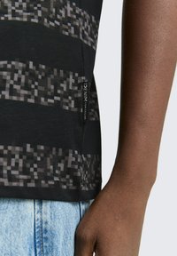 Zwarte korte mouwen shirt van textuurstof, voorzien van een gepixeld patroon in donkere tinten, met een etiket aan de zijkant en gedragen met denim.