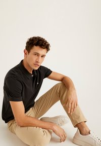 Černé polo triko s límečkem a krátkým rukávem; světle hnědé slim-fit kalhoty; bílé tenisky; model sedící s jednou nohou pokrčenou.