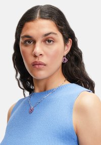 Ohrringe mit einem geometrischen pinken Design und einem metallischen Akzent, kombiniert mit einer silbernen Kettenhalskette. Das Model trägt ein blaues, geripptes, ärmelloses Oberteil.
