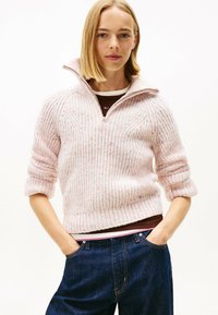 Femme jeune avec des cheveux blonds raides portant un pull zippé en tricot rose clair et un jean bleu denim foncé, les mains dans les poches.
