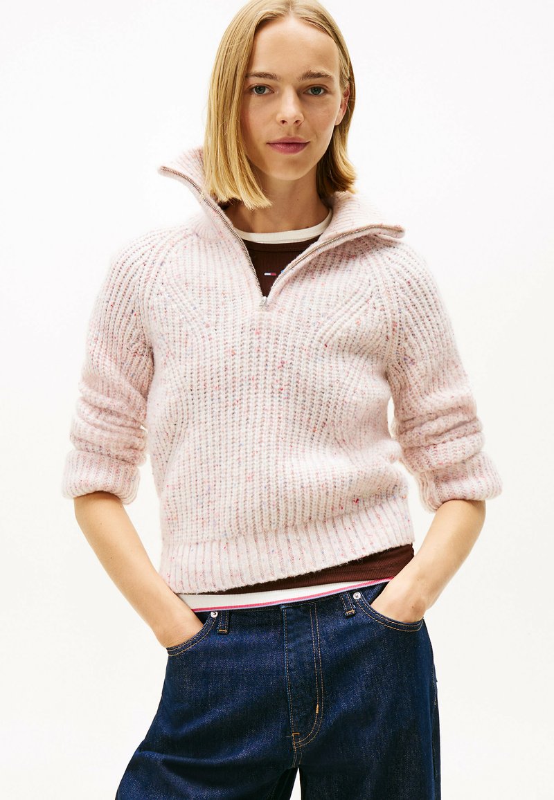 Femme jeune avec des cheveux blonds raides portant un pull zippé en tricot rose clair et un jean bleu denim foncé, les mains dans les poches.