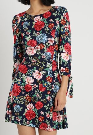 Frau trägt ein marineblaues Kleid mit roten, pinken und blauen Blumenmustern und dreiviertel Ärmeln, die an den Bündchen mit Schleifen gebunden sind.