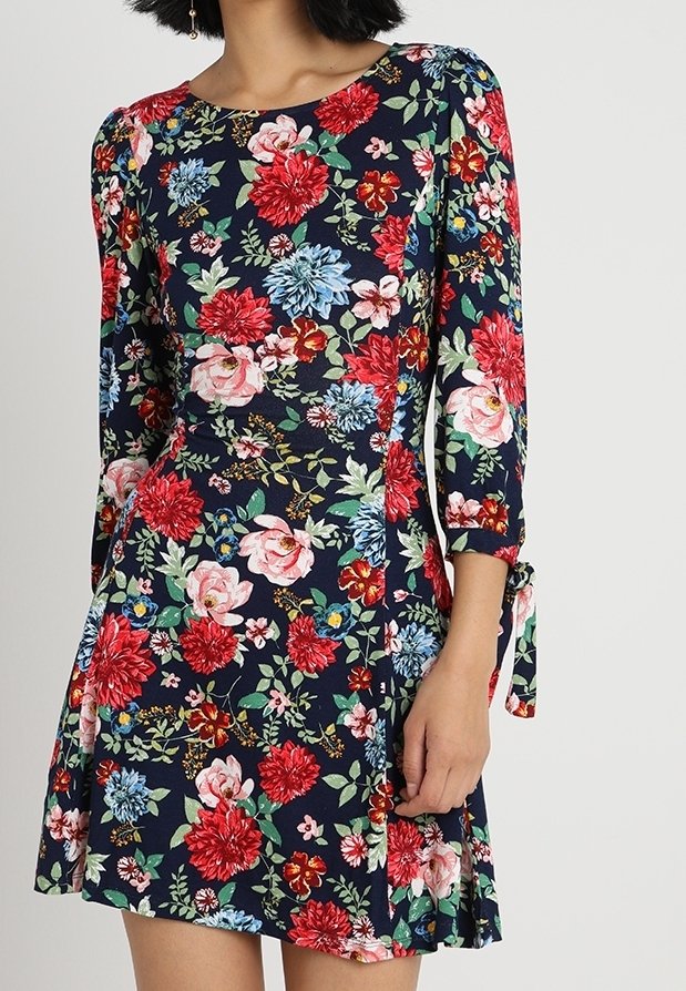 Femme portant une robe bleu marine avec des motifs floraux rouges, roses et bleus et des manches trois quarts nouées avec des nœuds aux poignets.