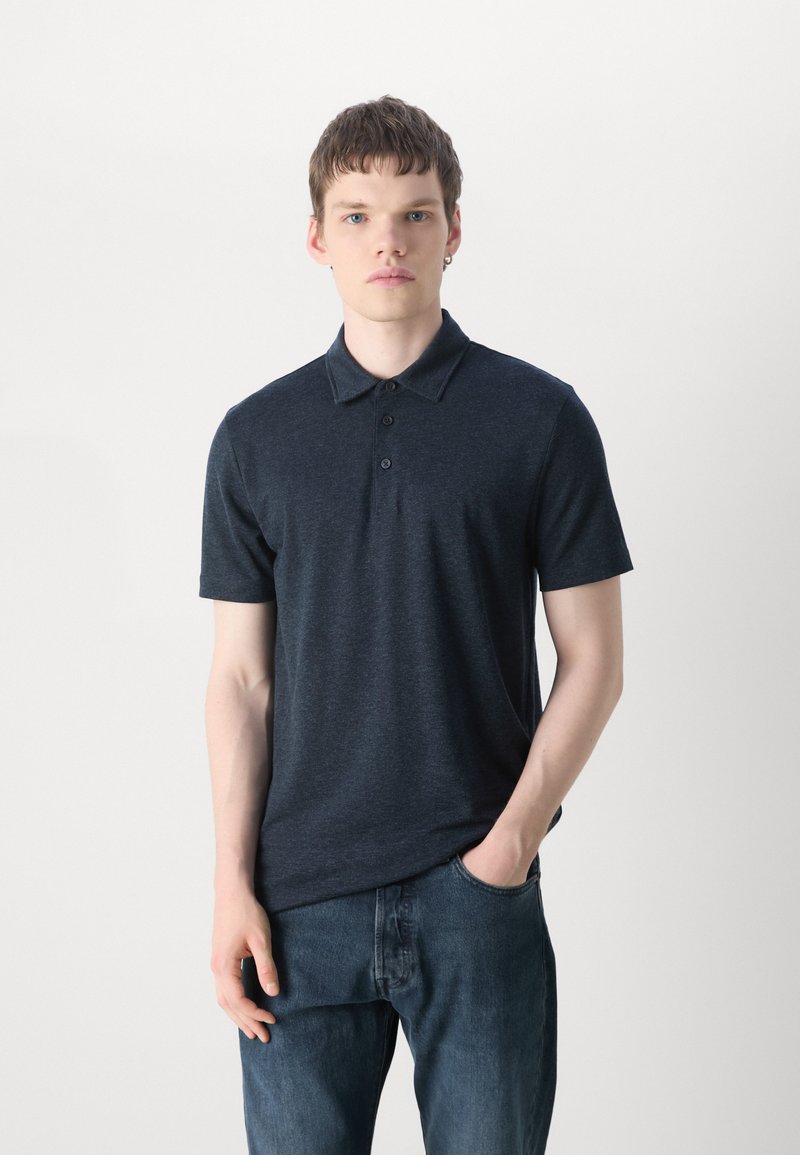 Marineblaue Polohemd aus weichem Stoff, mit einem kragenförmigen Hals und einer Knopfleiste mit drei Knöpfen. Kombiniert mit blauen Jeans, steht vor einem hellen Hintergrund.