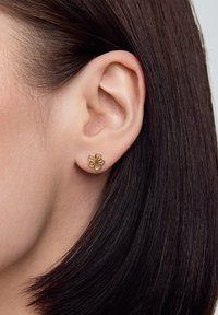 Złoty kolczyk w kształcie kwiatu z teksturowaną powierzchnią i centralnym elementem dekoracyjnym. Noszony na uchu modelki, w kontraście do ciemnych włosów.