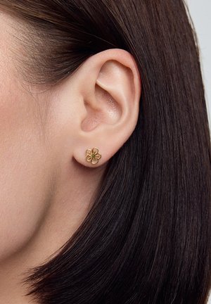 Złoty kolczyk w kształcie kwiatu z teksturowaną powierzchnią i centralnym elementem dekoracyjnym. Noszony na uchu modelki, w kontraście do ciemnych włosów.