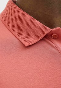 Korall színű polo póló, texturált hálós gallérral és sima testrésszel. A galléron gomb részlet található, valamint puha szövet textúrával rendelkezik.